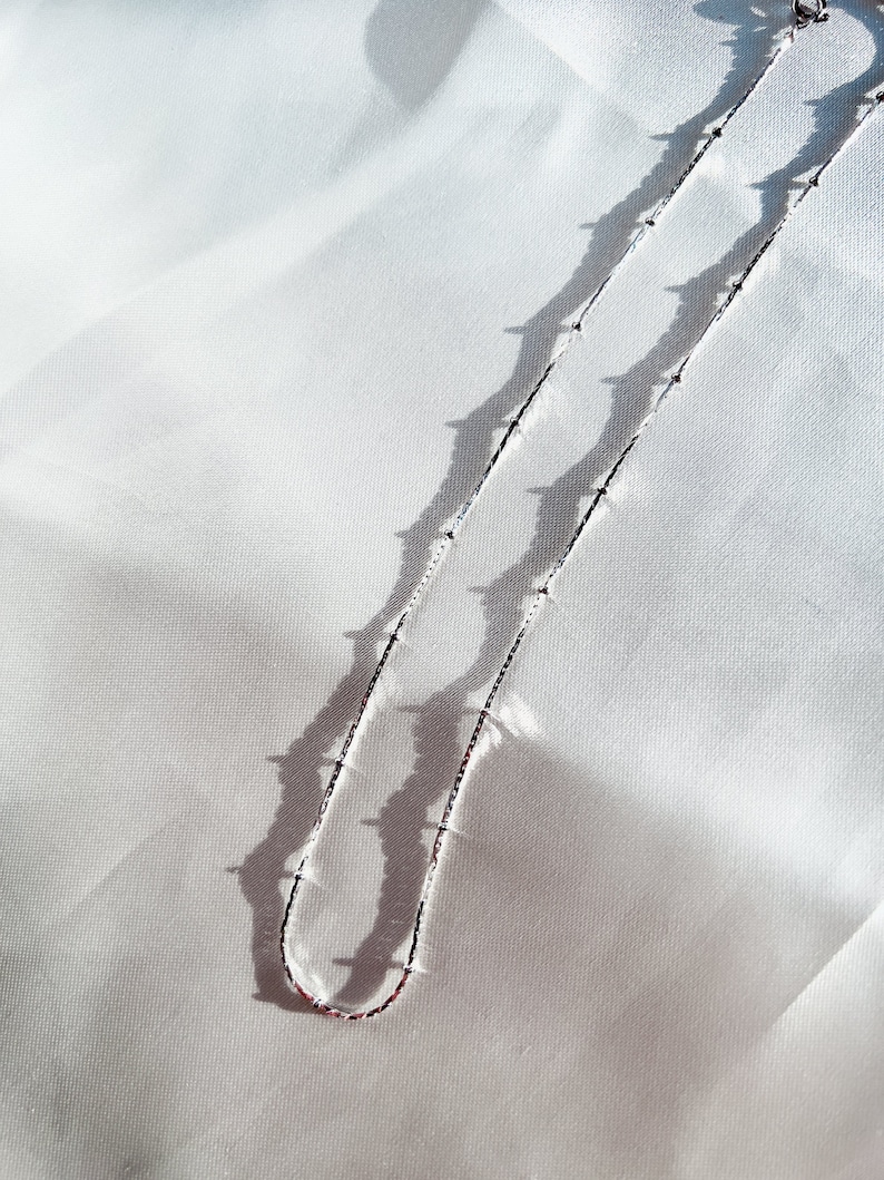 May include: A delicate silver necklace with small, evenly spaced beads. The necklace is displayed on a white, textured fabric, with shadows creating a contrasting effect. The chain is thin and elegant, suitable for everyday wear.