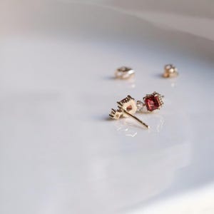 May include: A pair of gold-colored stud earrings with a square, deep red gemstone and a small, clear gemstone. The earrings are set on a white surface, with two earring backs in the background. The earrings have a delicate, ornate design.