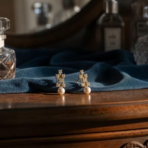May include: A pair of gold-toned stud earrings featuring a textured, rectangular metal frame with a small, round, white pearl dangling from the base of each piece. The earrings are displayed upright, showcasing the intricate, rough surface detail.