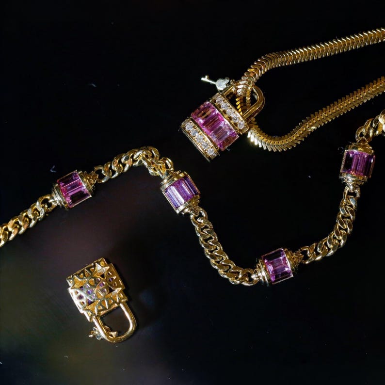 May include: A gold necklace with a lock pendant and a key, featuring pink rectangular gemstones and small diamonds. The necklace has a chain design with additional pink gemstones. The lock is open.