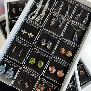 Op de afbeelding: Een witte lade-organizer met zwarte kaartjes waarop verschillende paar oorbellen worden getoond. De oorbellen zijn gemaakt van strass, parels en andere decoratieve elementen. De kaartjes hebben de tekst "BOITE LAQUE" en een website-adres.