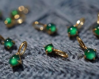 May include: Close-up of several gold-colored earrings with a small, round, vibrant green gemstone. The earrings are set against a textured, gray background, creating a contrast that highlights the jewelry's details.