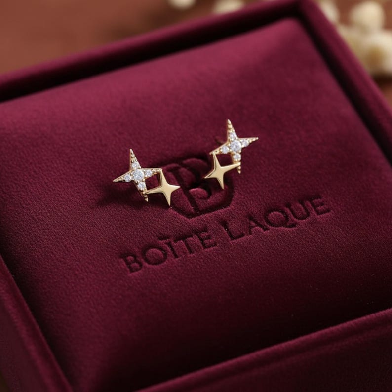 May include: Gold-coloured star stud earrings, each set with small, sparkling white gemstones. The earrings are presented in a burgundy velvet jewellery box. The box has the text "BOITE LAQUE" embossed on the inside.