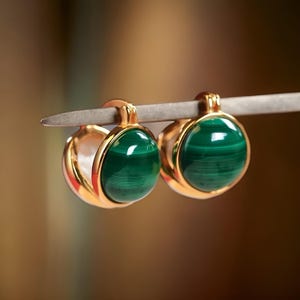 May include: A pair of gold hoop earrings with green malachite stones. The earrings are on a silver stand.