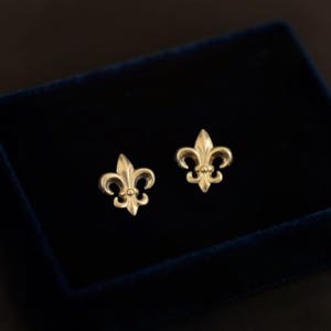 K&ouml;nnte beinhalten: Ein Paar goldfarbene Fleur-de-Lis-Ohrstecker. Die Ohrringe werden auf einer dunkelblauen Samtoberfl&auml;che pr&auml;sentiert. Die Ohrringe haben ein detailliertes, dreibl&auml;ttriges Design mit einem kleinen, runden Akzent an der Basis jedes Blattes.