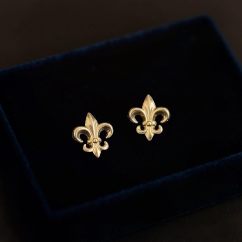 K&ouml;nnte beinhalten: Ein Paar goldfarbene Fleur-de-Lis-Ohrstecker. Die Ohrringe werden auf einer dunkelblauen Samtoberfl&auml;che pr&auml;sentiert. Die Ohrringe haben ein detailliertes, dreibl&auml;ttriges Design mit einem kleinen, runden Akzent an der Basis jedes Blattes.