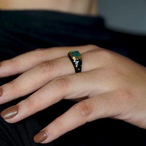 May include: A black ring with a green gemstone and gold accents. The ring is worn on a person's finger.