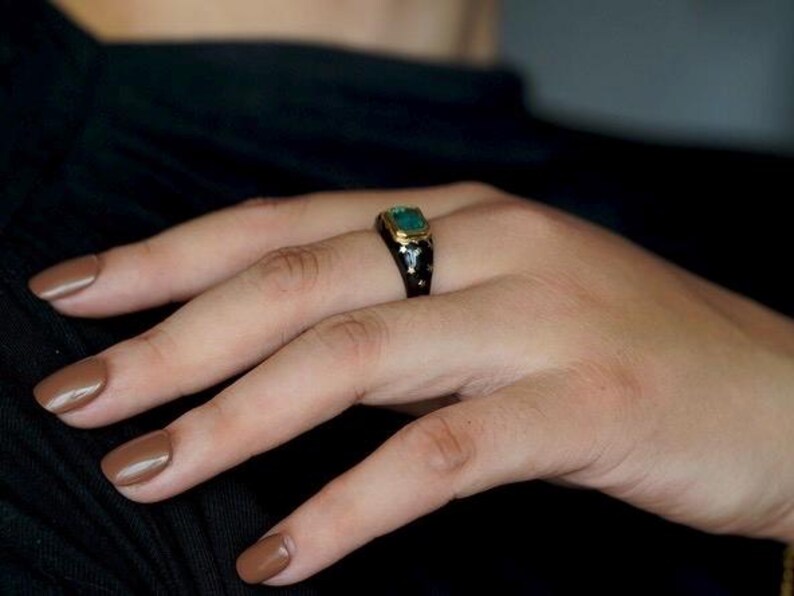 May include: A black ring with a green gemstone and gold accents. The ring is worn on a person's finger.
