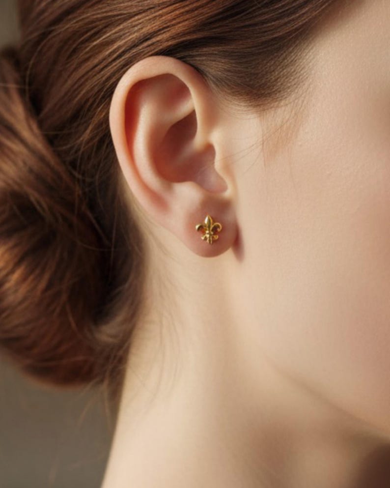 K&ouml;nnte beinhalten: Ein goldfarbener Ohrstecker in Form einer Fleur-de-Lis ist an einem Ohr zu sehen. Der Ohrring ist klein und detailliert, mit einem klassischen dreibl&auml;ttrigen Design. Der Hintergrund ist unscharf, wodurch der Fokus auf dem Schmuck liegt.
