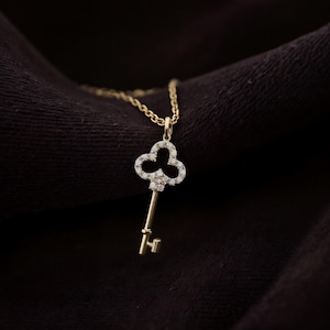 May include: A gold-colored key pendant necklace. The key design features a clover-shaped top encrusted with small, sparkling stones. The key's stem and the chain are also gold-colored, set against a dark background.
