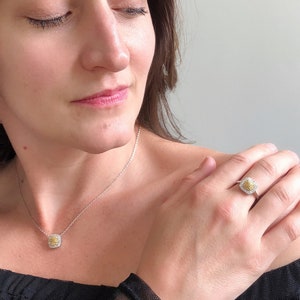 Puede incluir: Una mujer lleva una blusa negra con un delicado collar de cadena plateada y un peque&ntilde;o colgante de diamante amarillo. Tambi&eacute;n lleva un anillo con un gran diamante amarillo rodeado de diamantes m&aacute;s peque&ntilde;os en su dedo anular.