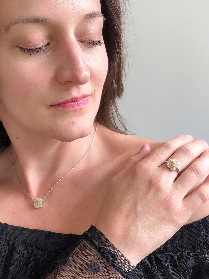 Puede incluir: Una mujer lleva una blusa negra con un delicado collar de cadena plateada y un peque&ntilde;o colgante de diamante amarillo. Tambi&eacute;n lleva un anillo con un gran diamante amarillo rodeado de diamantes m&aacute;s peque&ntilde;os en su dedo anular.