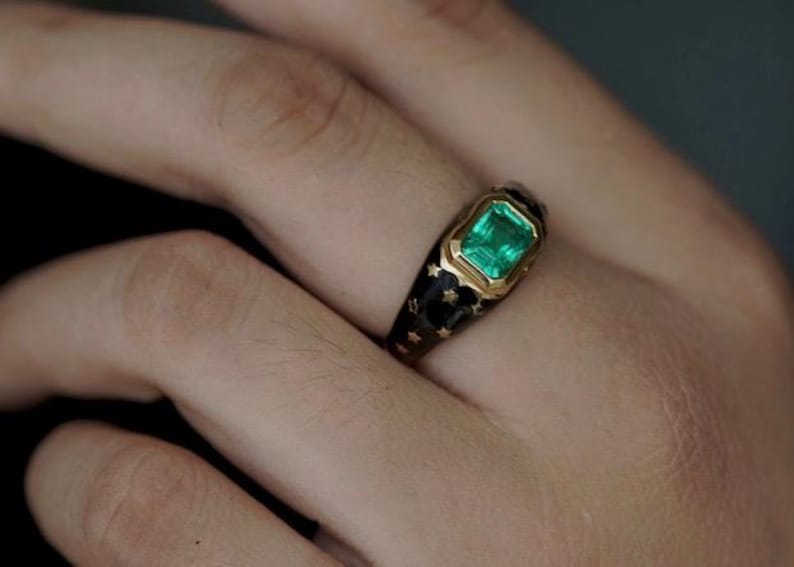 May include: A gold ring with a green emerald gemstone set in a black enamel band with gold stars.