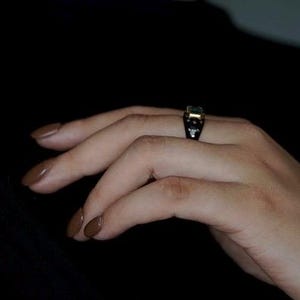 May include: A hand wearing a ring with a green gemstone and black and gold accents.