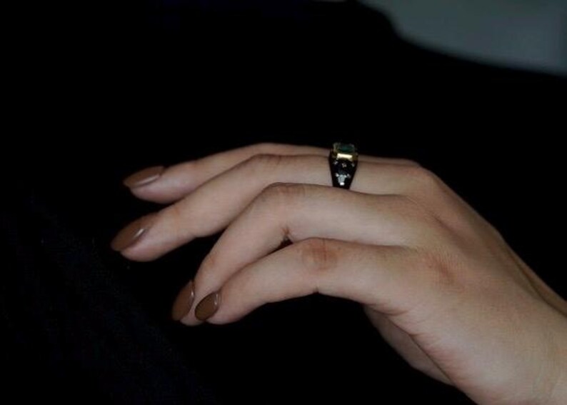 May include: A hand wearing a ring with a green gemstone and black and gold accents.