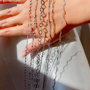 May include: A collection of delicate silver necklaces with various chain designs. The necklaces are displayed against a soft, white background, showcasing their intricate patterns and reflective surfaces. The chains include flat, geometric, and wavy styles.