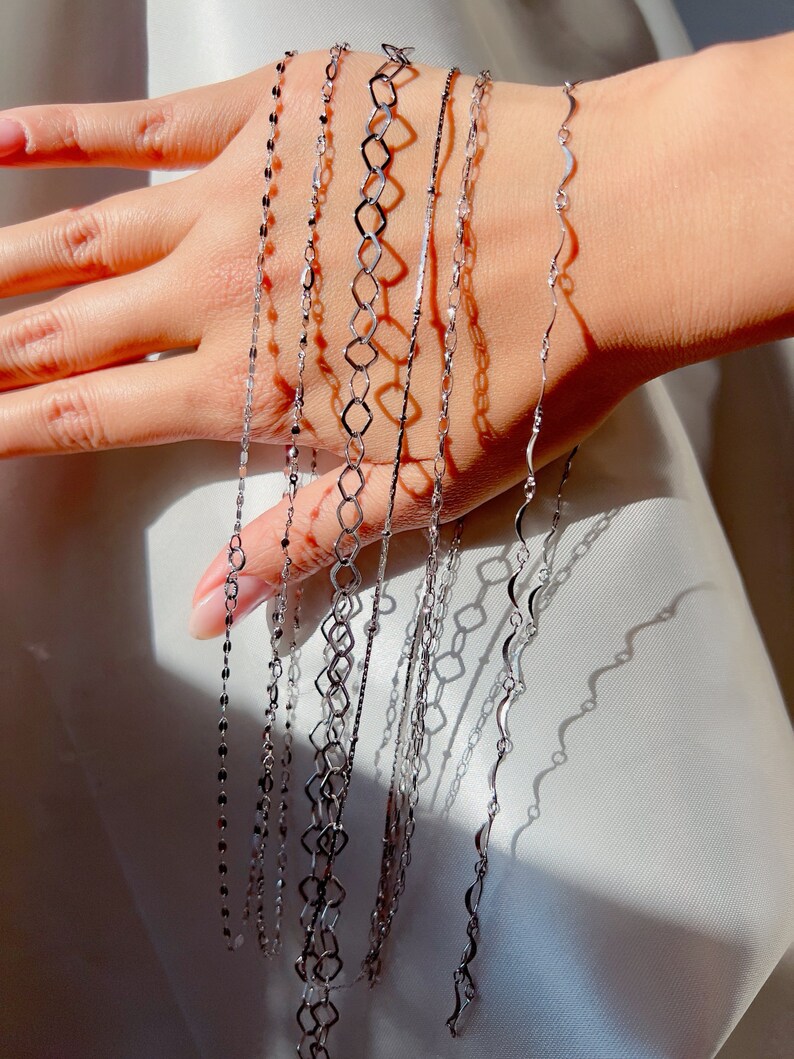 May include: A collection of delicate silver necklaces with various chain designs. The necklaces are displayed against a soft, white background, showcasing their intricate patterns and reflective surfaces. The chains include flat, geometric, and wavy styles.