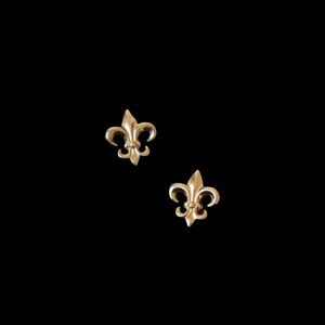 K&ouml;nnte beinhalten: Zwei goldfarbene Fleur-de-Lis-Ohrstecker. Jeder Ohrring hat ein stilisiertes Design mit drei Bl&uuml;tenbl&auml;ttern und einem zentralen Element. Die Ohrringe sind vor einem schwarzen Hintergrund platziert, der ihre filigranen Details und ihren metallischen Glanz hervorhebt.