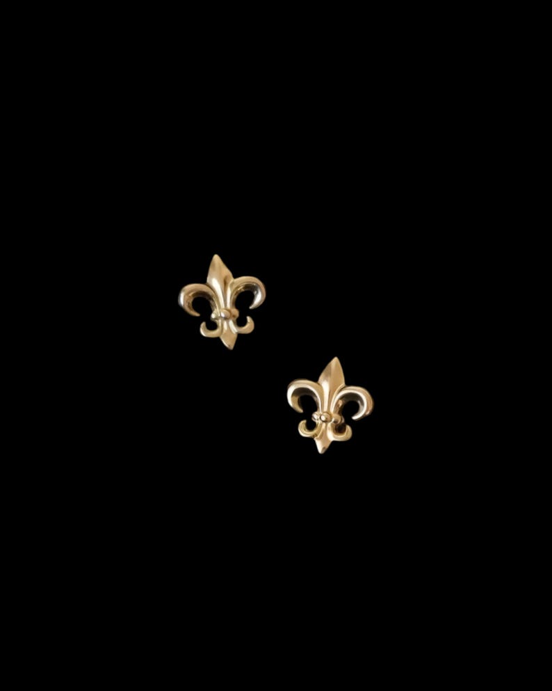 K&ouml;nnte beinhalten: Zwei goldfarbene Fleur-de-Lis-Ohrstecker. Jeder Ohrring hat ein stilisiertes Design mit drei Bl&uuml;tenbl&auml;ttern und einem zentralen Element. Die Ohrringe sind vor einem schwarzen Hintergrund platziert, der ihre filigranen Details und ihren metallischen Glanz hervorhebt.