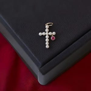 May include: A white pearl cross pendant with a small red gemstone hanging from the bottom. The pendant is on a black background.