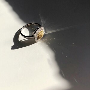 Puede incluir: Un anillo de plata con una piedra central de forma cuadrada. La piedra central es un diamante amarillo rodeado de diamantes blancos m&aacute;s peque&ntilde;os.