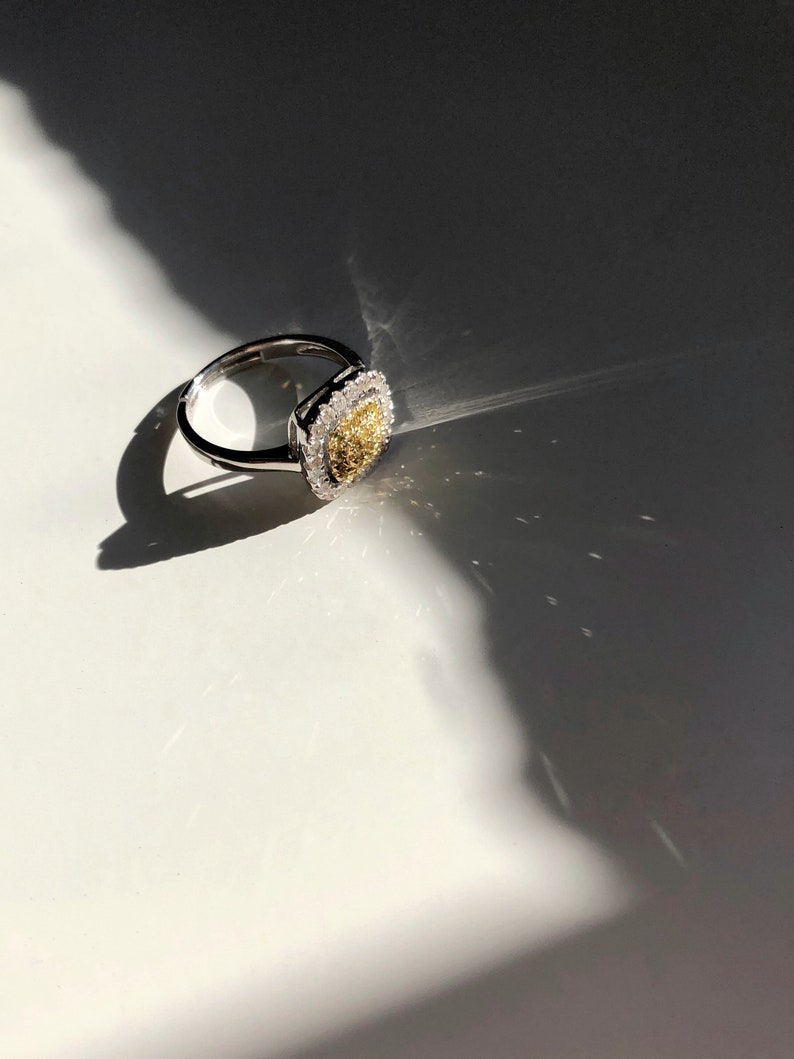 Puede incluir: Un anillo de plata con una piedra central de forma cuadrada. La piedra central es un diamante amarillo rodeado de diamantes blancos m&aacute;s peque&ntilde;os.
