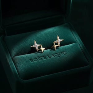 May include: Gold-coloured star earrings, each with small, sparkling stones, presented in a dark green velvet jewellery box. The box is embossed with the text "BOITE LAQUE" on the front.
