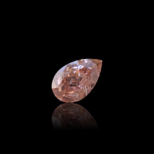 May include: A pear-shaped, light brown diamond with a brilliant cut. The gemstone is set against a black background, with a mirrored reflection below. The diamond's facets catch the light, showcasing its clarity and color.
