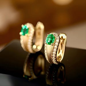 May include: A pair of gold hoop earrings, each featuring a vibrant green emerald center stone surrounded by a halo of small, sparkling diamonds. The earrings have a brushed gold finish and are displayed on a reflective black surface.