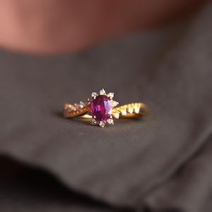 May include: A gold ring with a central oval ruby, encircled by small diamonds. The band is also set with diamonds. The ring's design creates a halo effect around the ruby. The ring is displayed against a dark backdrop.