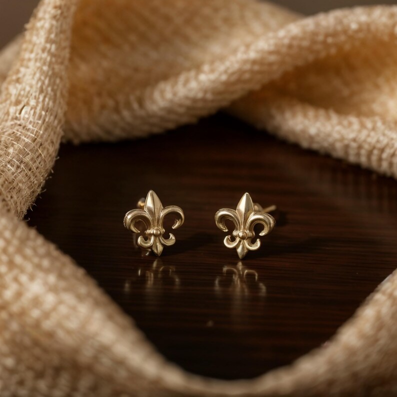K&ouml;nnte beinhalten: Ein Paar goldfarbene Fleur-de-Lis-Ohrstecker. Die Ohrringe werden auf einer dunkelbraunen Oberfl&auml;che mit einem beigefarbenen Stoff im Hintergrund pr&auml;sentiert. Die Ohrringe sind klein und detailliert.