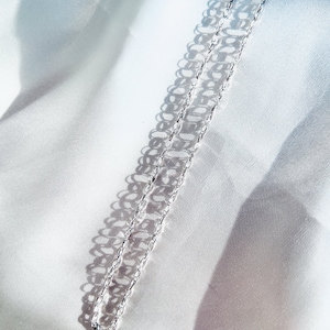 May include: A delicate silver chain necklace with a repeating oval link design. The necklace is displayed on a white satin fabric, with shadows creating a pattern. The chain is a simple, elegant piece of jewelry.