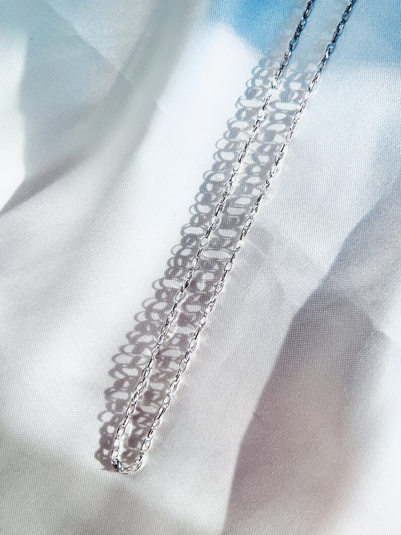 May include: A delicate silver chain necklace with a repeating oval link design. The necklace is displayed on a white satin fabric, with shadows creating a pattern. The chain is a simple, elegant piece of jewelry.