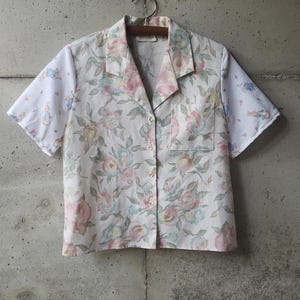 May include: A short-sleeved button-down shirt with a floral pattern in pastel pink, blue, and yellow. The sleeves have a white background with a cartoon rabbit design. The shirt is hanging on a wooden hanger against a gray concrete wall.