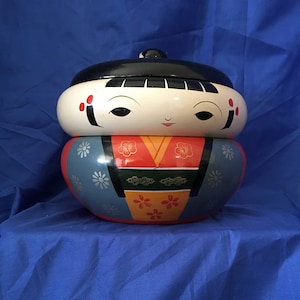 Op de afbeelding: Een traditionele Japanse nestpop, of 'kokeshi', met een geschilderd gezicht en bloemmotieven. De pop is gemaakt van hout en heeft een blauw, rood en geel kleurenschema.