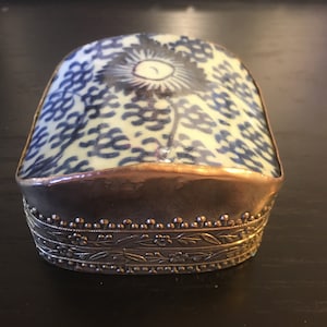 Puede incluir: Una pequeña caja rectangular con un diseño floral azul y blanco en la parte superior. La caja tiene un borde de metal plateado alrededor de los bordes y un patrón floral decorativo en la parte inferior.