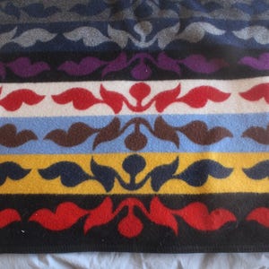 Può includere: Una coperta colorata con un motivo floreale ripetuto. La coperta presenta strisce orizzontali in vari colori, tra cui grigio, blu navy, viola, bianco, rosso, azzurro, marrone, giallo e nero. Il motivo floreale è un motivo stilizzato di foglie e steli.