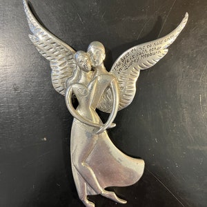Puede incluir: Una figurita de ángel de plata con las alas extendidas, sosteniendo un objeto con forma de corazón. El ángel es una pareja que se abraza, y la cita "Lo mejor a lo que aferrarse en la vida es el uno al otro" de Audrey Hepburn está inscrita en las alas.