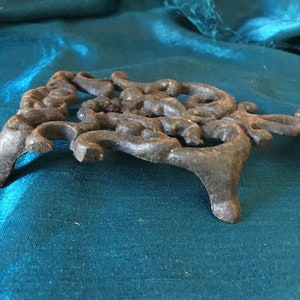 May include: A dark, ornate cast iron trivet with a decorative, openwork design. The trivet has a three-legged base and is designed to protect surfaces from hot cookware. The color is a dark, aged black.