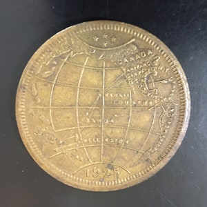 Puede incluir: Una moneda de color bronce con un mapa del mundo. La moneda tiene el año 1925 inscrito en ella. El mapa muestra las ubicaciones de Canadá, San Francisco, Hawái, Samoa, Wellington y América del Sur.