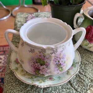 Könnte beinhalten: Eine weiße Porzellan-Zuckerdose mit Blumenmuster und einer passenden Untertasse. Die Schale hat einen Deckel und zwei Griffe. Das Blumenmuster zeigt rosa und violette Blumen mit grünen Blättern.