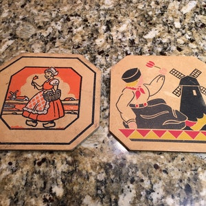 May include: Two vintage Dutch-themed ceramic tiles. One tile features a woman in traditional Dutch clothing holding a flower and a basket. The other tile features a man in traditional Dutch clothing sitting near a windmill.
