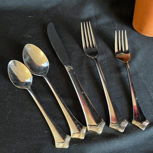 Könnte beinhalten: Ein Set aus Edelstahl-Besteck, darunter zwei Löffel, ein Messer und zwei Gabeln. Das Besteck hat ein dekoratives goldfarbenes Muster an der Basis der Griffe. Die Artikel sind auf einer dunklen Oberfläche angeordnet.