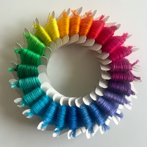 May include: A rainbow assortment of embroidery floss wound on white plastic bobbins. The floss is arranged in a circle, with the colors transitioning from red to orange, yellow, green, blue, and purple.