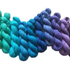 May include: A collection of ten twisted skeins of yarn arranged in a horizontal gradient. The colors transition from bright turquoise and teal on the left, through shades of royal blue, to deep violet and purple on the right side of the display.