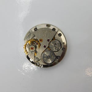 Könnte beinhalten: Ein silbernes Uhrwerk mit einem goldfarbenen Aufzugstift und Zahnrädern. Das Werk ist mit "Zarya" beschriftet und hat einen roten Stein.