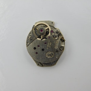Könnte beinhalten: Nahaufnahme eines goldfarbenen Uhrwerks mit der Aufschrift "17 Jewels". Das Uhrwerk ist freigelegt und zeigt die inneren Funktionsteile der Uhr.