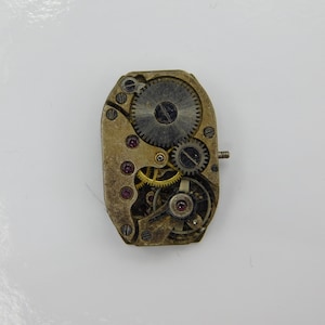 Könnte beinhalten: Nahaufnahme der Rückseite eines Vintage-Uhrwerks mit goldfarbenen Zahnrädern und einer silberfarbenen Platte. Das Uhrwerk ist freigelegt und zeigt die komplizierten Funktionsweisen der Uhr.