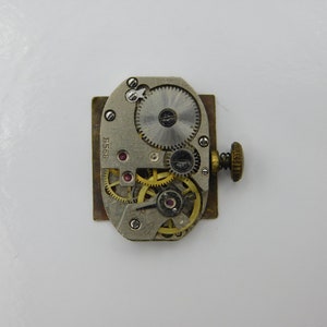 Op de afbeelding: Een zilveren en gouden uurwerk met een reeks tandwielen en een opwindas. Het uurwerk is gemarkeerd met "8568".