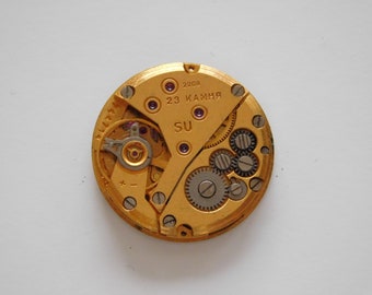 May include: A gold-colored watch movement with a silver-colored gear system. The movement has the text "2209 23 КАМНЯ SU" printed on it.